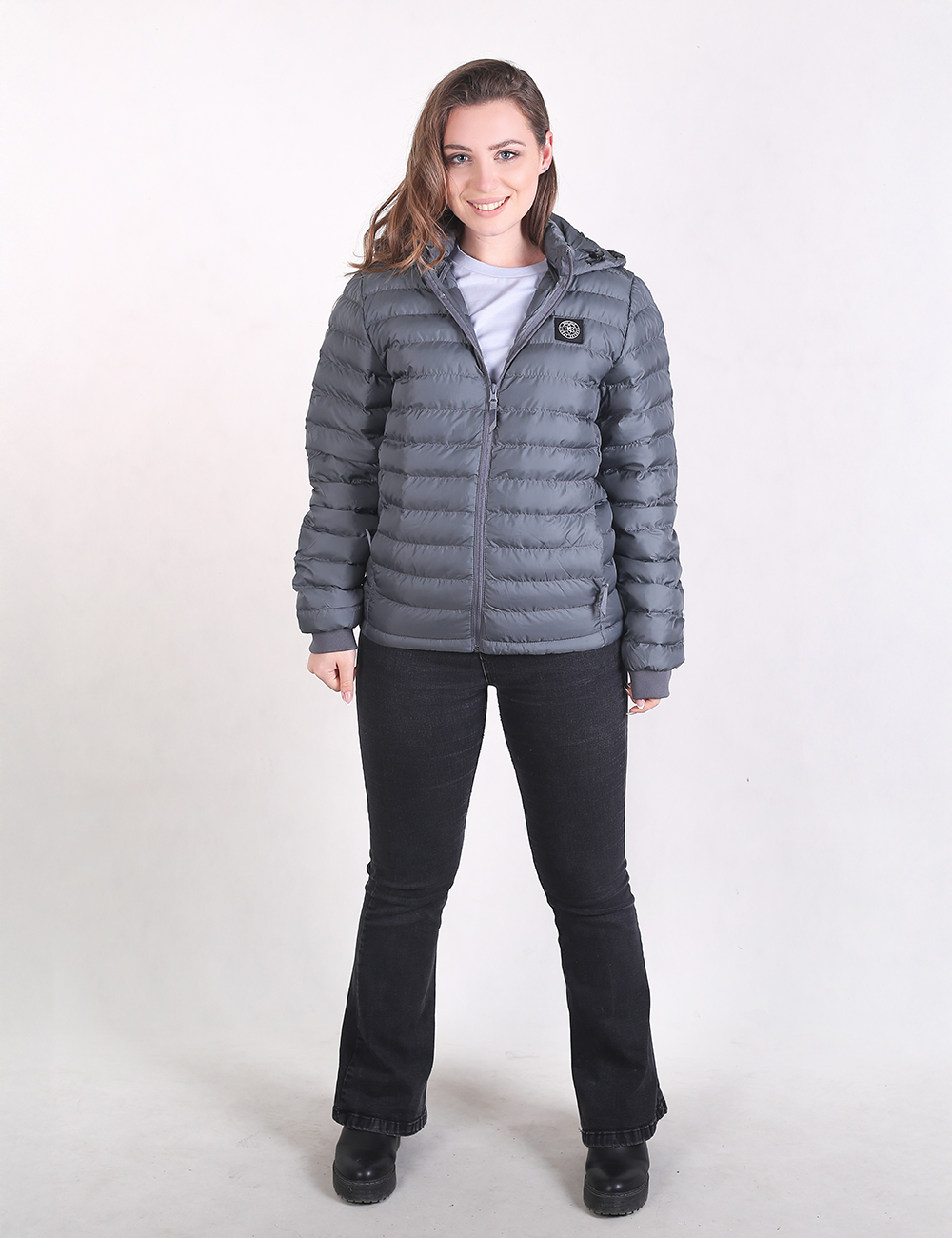 A woman modeling a gray hooded puffer jacket with horizontal quilting, paired with black jeans and black shoes, standing against a plain white studio background.