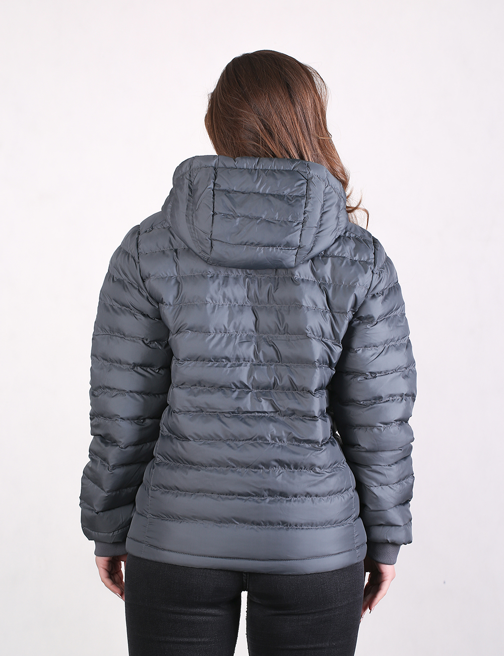A woman is viewed from behind, wearing a dark gray quilted puffer jacket with horizontal baffles and an attached hood, paired with black pants against a plain, light-colored background.