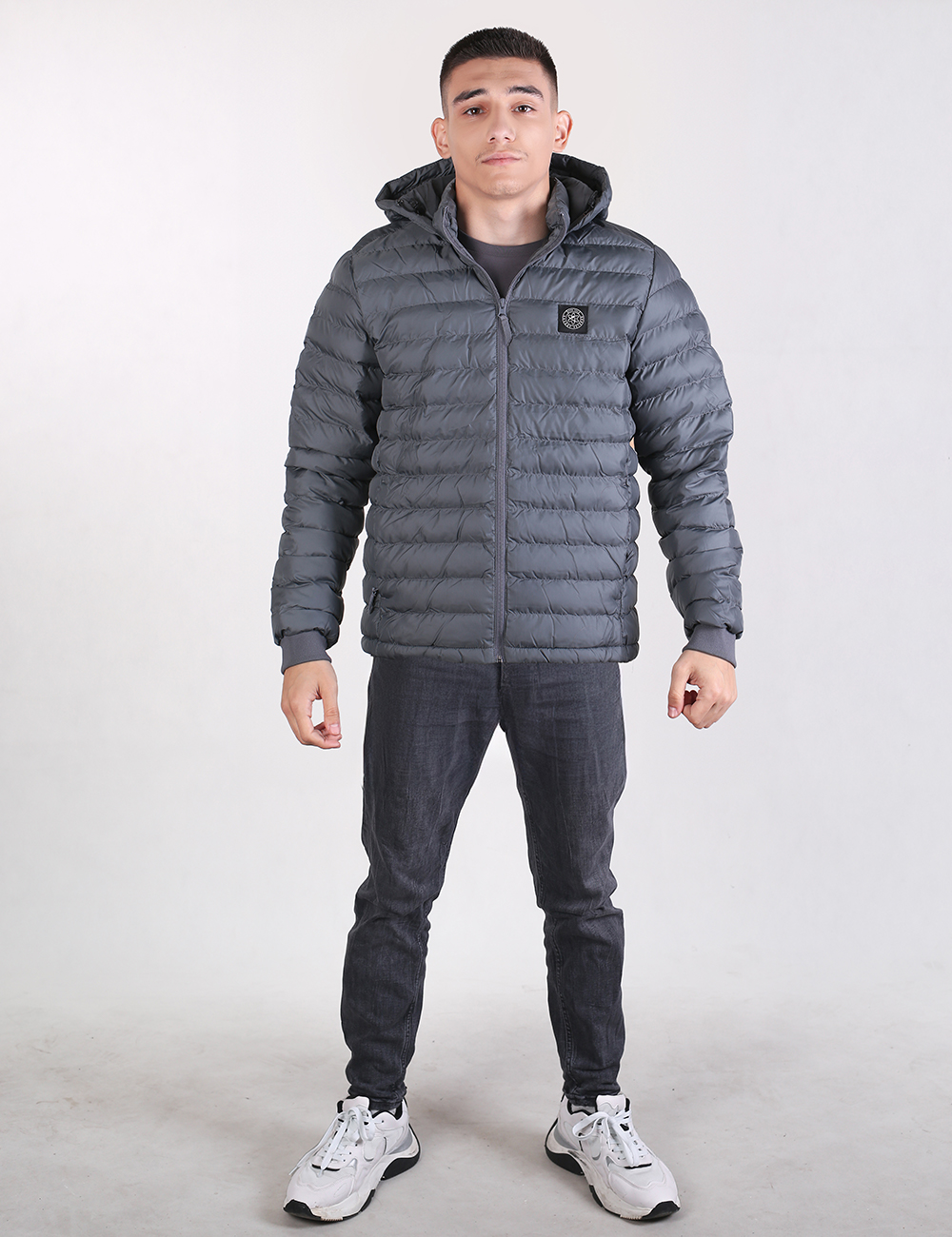 A young man stands facing forward against a plain white background, wearing a dark grey hooded puffer jacket with horizontal quilting and a square logo on the left chest. He is also wearing dark charcoal grey slim-fit jeans and white chunky athletic sneakers.