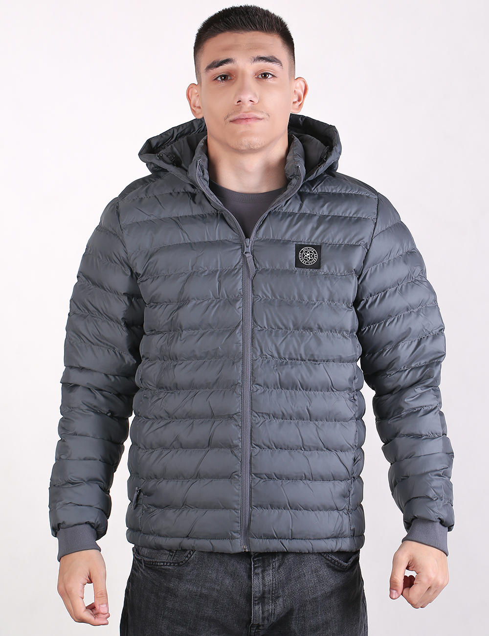 A man wearing a dark gray hooded puffer jacket with horizontal quilting, a front zipper, and a circular chest logo patch, paired with dark gray jeans, against a plain light background.
