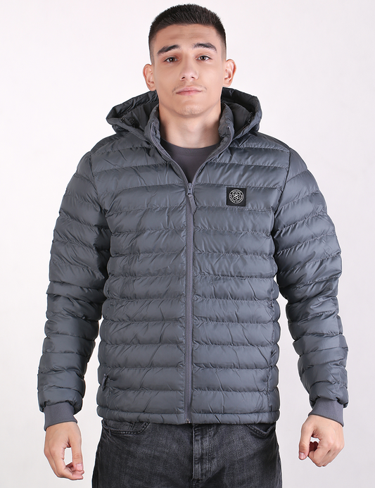 A man wearing a dark gray hooded puffer jacket with horizontal quilting, a front zipper, and a circular chest logo patch, paired with dark gray jeans, against a plain light background.