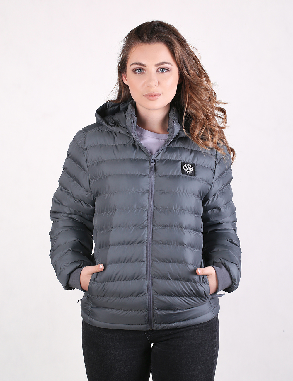 A young woman with long, wavy brown hair is standing and looking directly forward while modeling a dark grey, hooded puffer jacket. The jacket features horizontal quilted panels, a front zipper closure, a small circular logo patch on the left chest, and side pockets where she has placed her hands. She is wearing black pants, and the background is a solid, light off-white color.