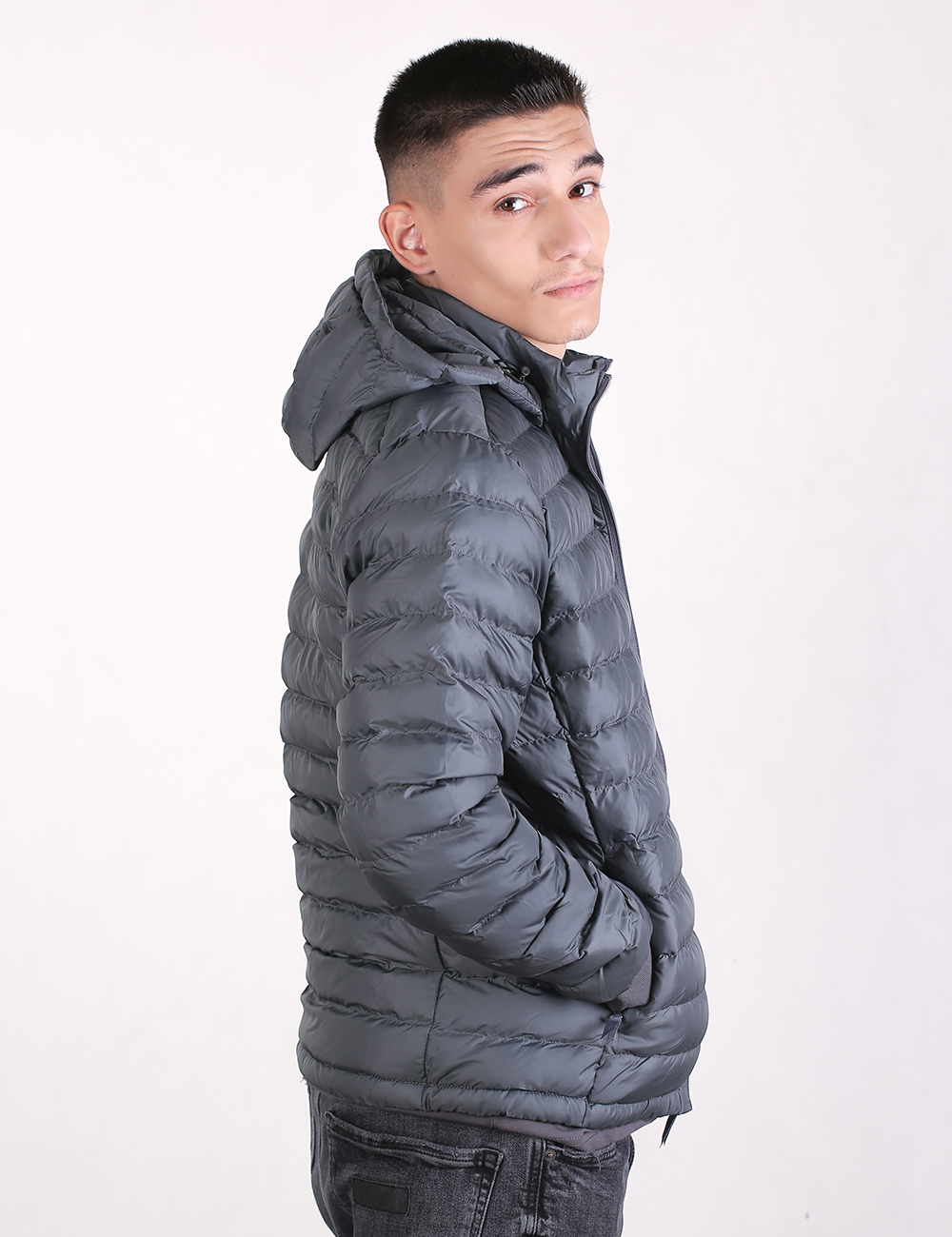 A man with a short buzz cut haircut stands in a side-profile view against a plain, off-white background. He is turned slightly toward the camera, looking over his right shoulder. He wears a grey puffer jacket with thin, horizontal quilting and an attached hood, paired with dark grey or black jeans. His right hand is tucked into the jacket pocket.