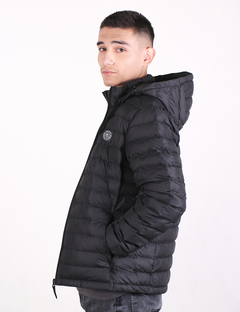 A side-profile shot of a young man wearing a black lightweight padded puffer jacket with a hood and a circular logo patch on the chest. He has short dark hair and is looking directly back over his shoulder against a plain white background.