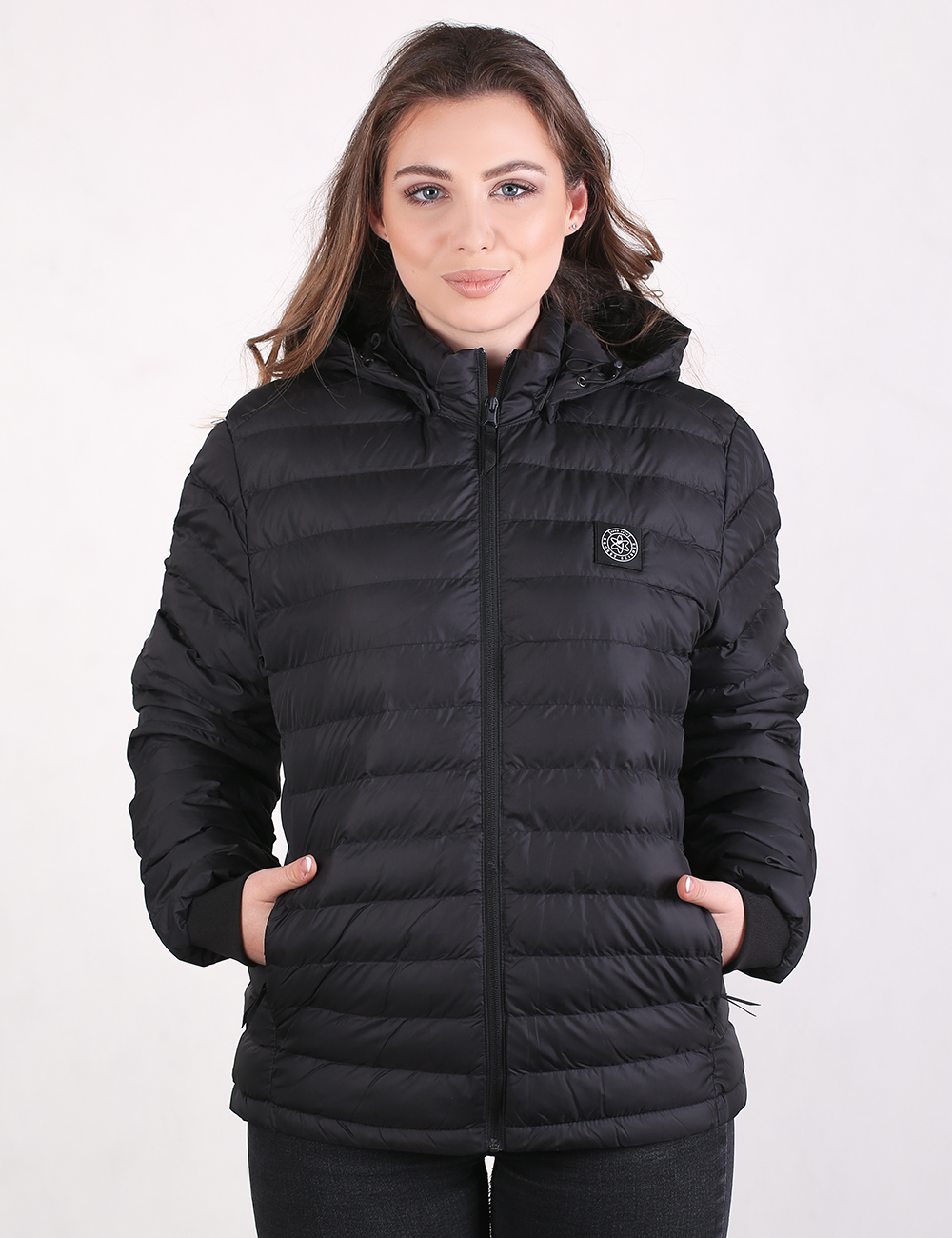 A woman modeling a black hooded puffer jacket with horizontal quilting, a front zipper, and a small circular chest logo, photographed against a plain light gray studio background.