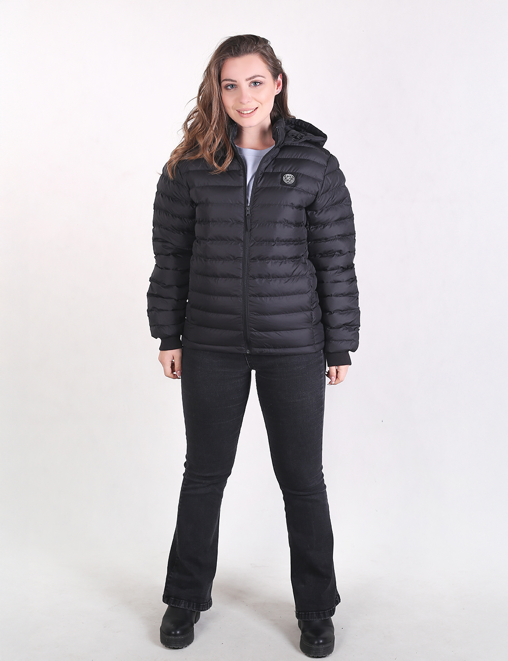 A woman modeling a black hooded puffer jacket with horizontal quilting, paired with black jeans and black shoes, against a plain white studio background.