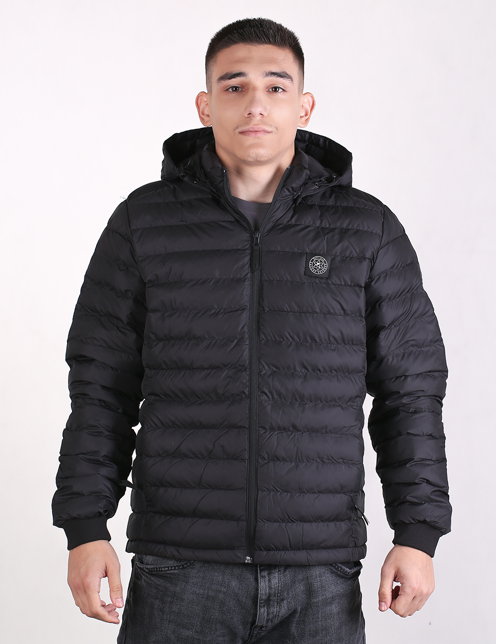 A medium-shot photograph of a young man with a short haircut, wearing a black quilted bubble puffer jacket with a hood and zipped up to the neck. The jacket has an elasticated cuff at the wrist and a circular logo badge on the left chest. He is facing forward against a plain white background, wearing dark gray jeans.