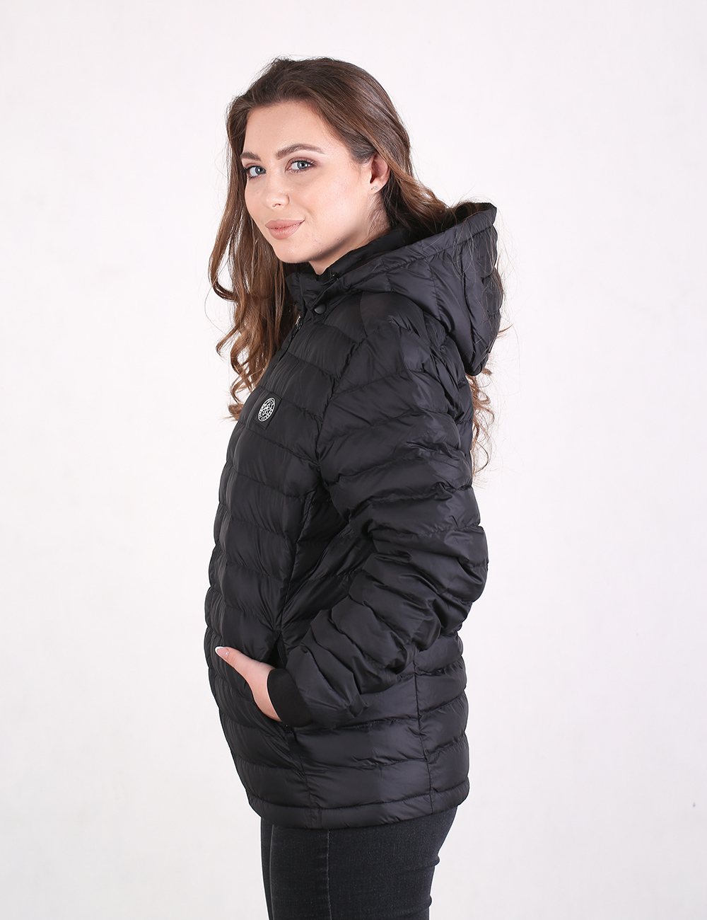 A woman wearing a black hooded puffer jacket, shown in profile against a plain white studio background.