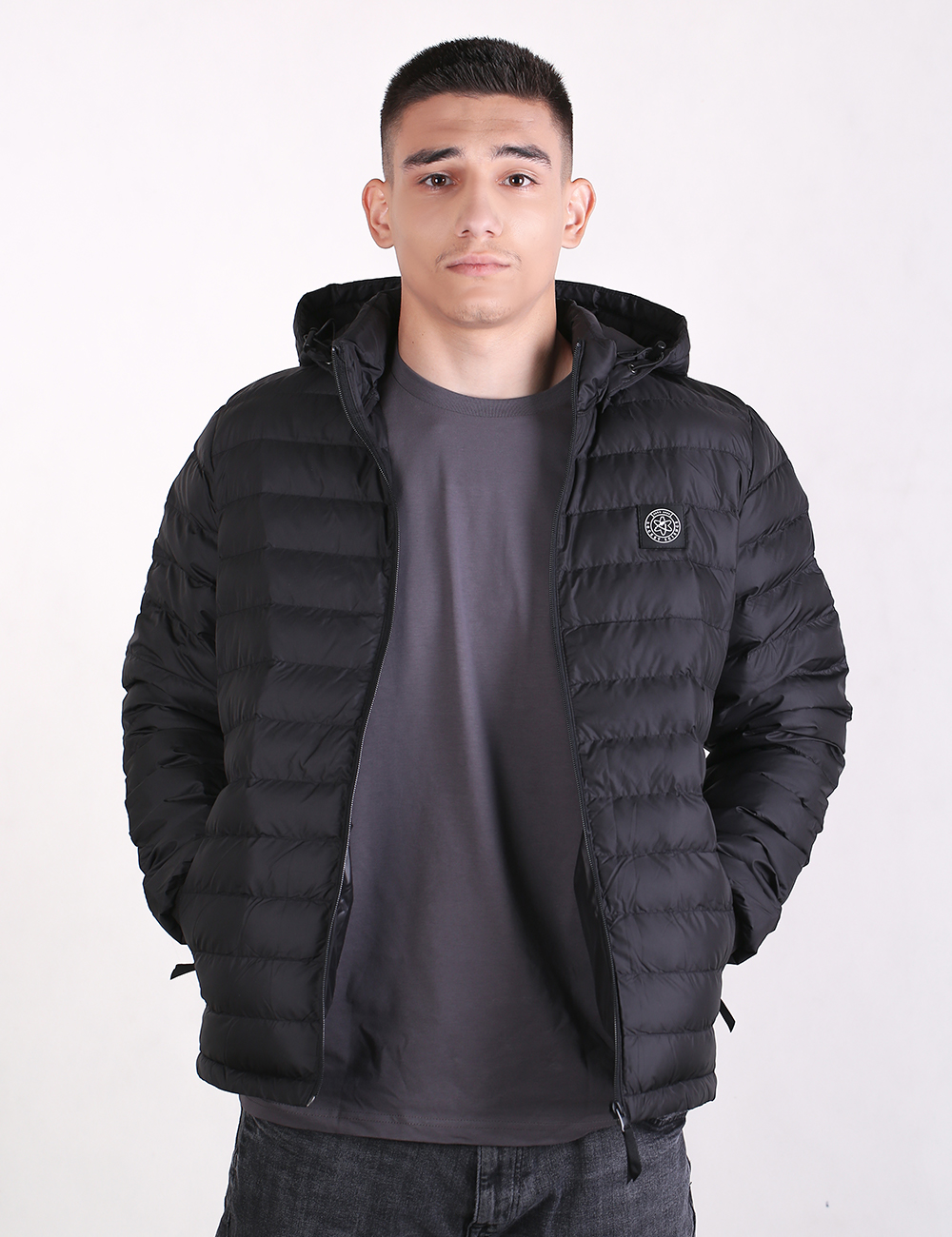 A man wearing a black hooded puffer jacket over a dark gray t-shirt and jeans, standing against a plain white studio background.