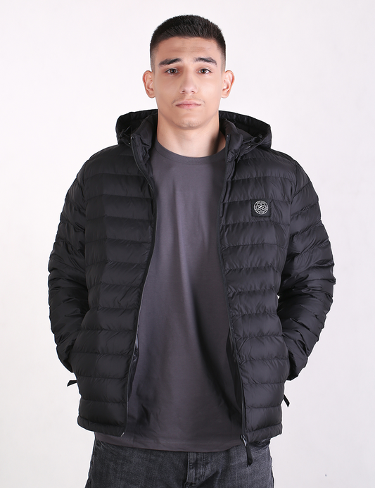 A man wearing a black hooded puffer jacket over a dark gray t-shirt and jeans, standing against a plain white studio background.