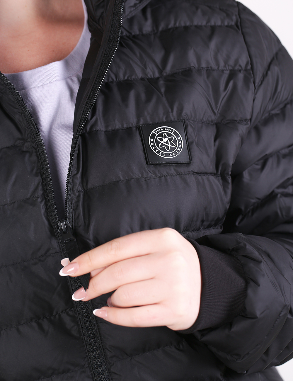 A close-up shot shows a person wearing a black quilted puffer jacket over a white shirt. The person's left hand, featuring long manicured nails, rests gently near the main front zipper. On the left chest area of the jacket, there is a small, square black patch containing a white circular atomic-style logo with text reading "ROCKET SCIENCE".