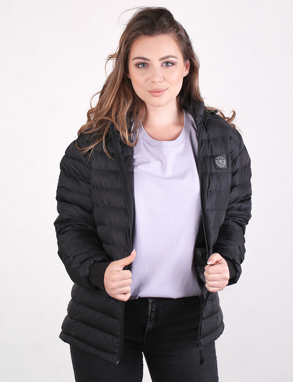 A woman wearing a black puffer jacket over a light purple t-shirt and black jeans, standing against a plain white studio background.
