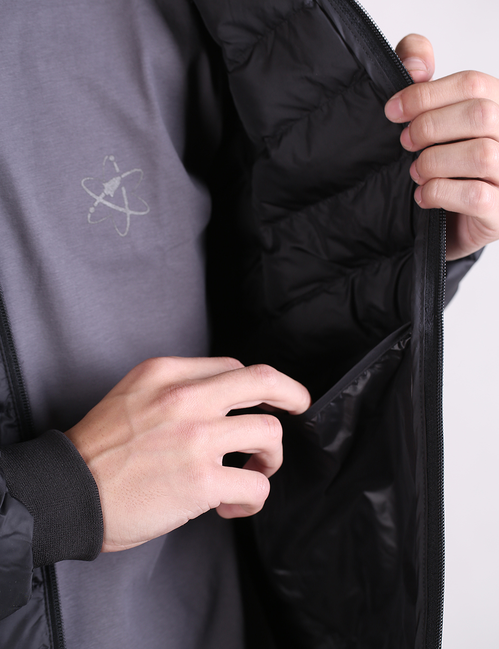 Close-up of a person wearing a black puffer jacket over a gray shirt, opening the jacket to show the interior lining and zipper pocket.