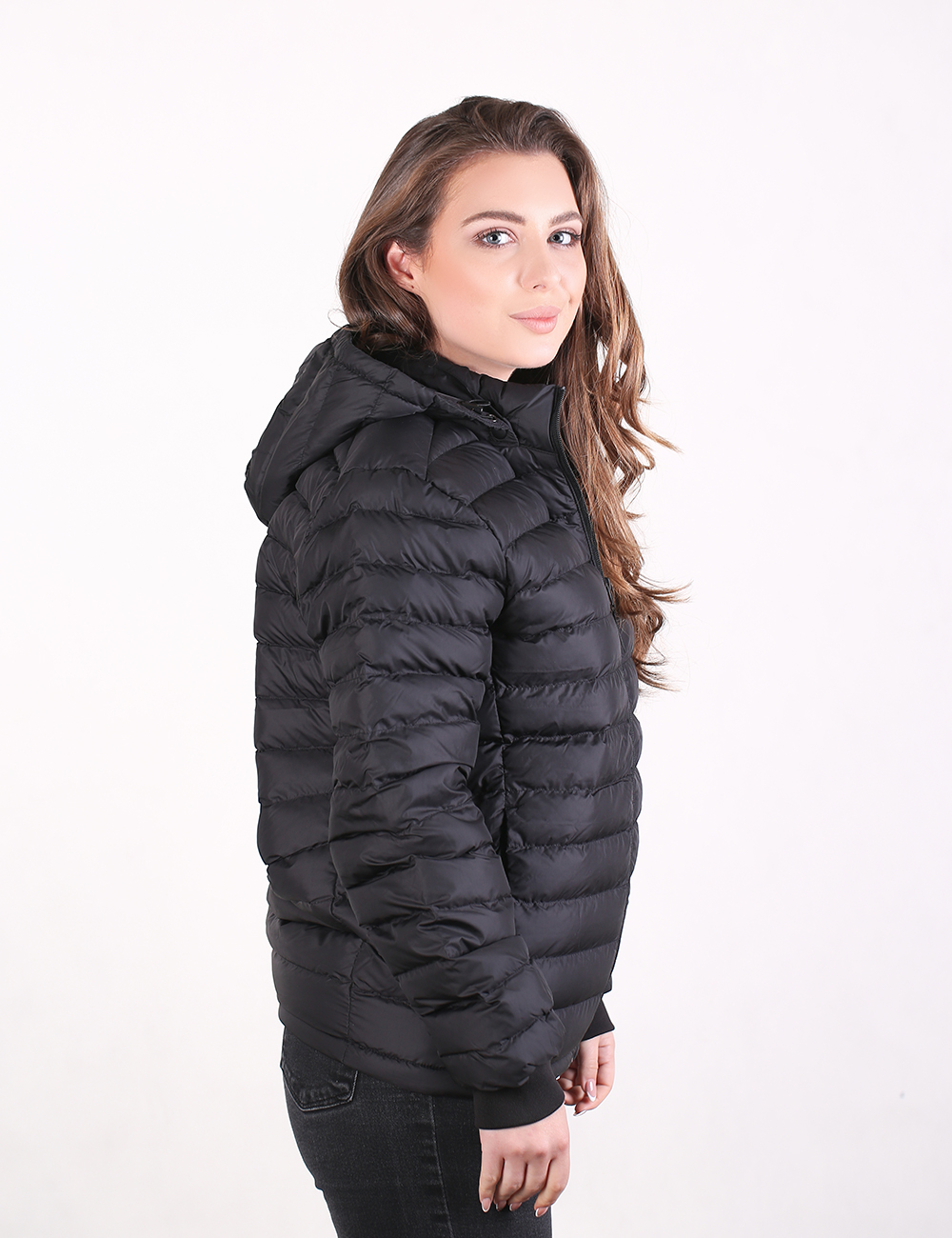 A woman wearing a black hooded puffer jacket, shown in profile against a plain white studio background.