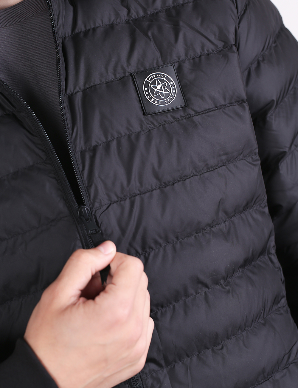 A close-up shot focuses on a black Rocket Science padded jacket. A hand is shown pulling up the front zipper. On the left chest area, there is a prominent, circular black patch with white text and an atom logo design that reads "ROCKET SCIENCE".
