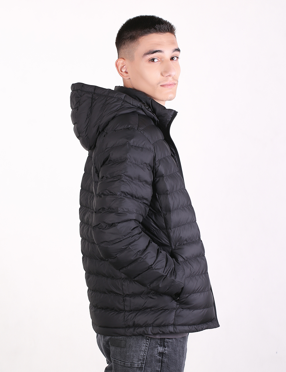 A young man with short, dark hair is shown from the side, looking back over his shoulder toward the camera against a plain white background. He is wearing a black, quilted puffer jacket with an attached hood and dark grey jeans.