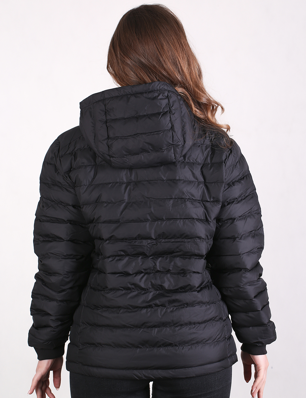A woman viewed from behind wearing a black hooded puffer jacket with horizontal quilting, set against a plain white studio background.
