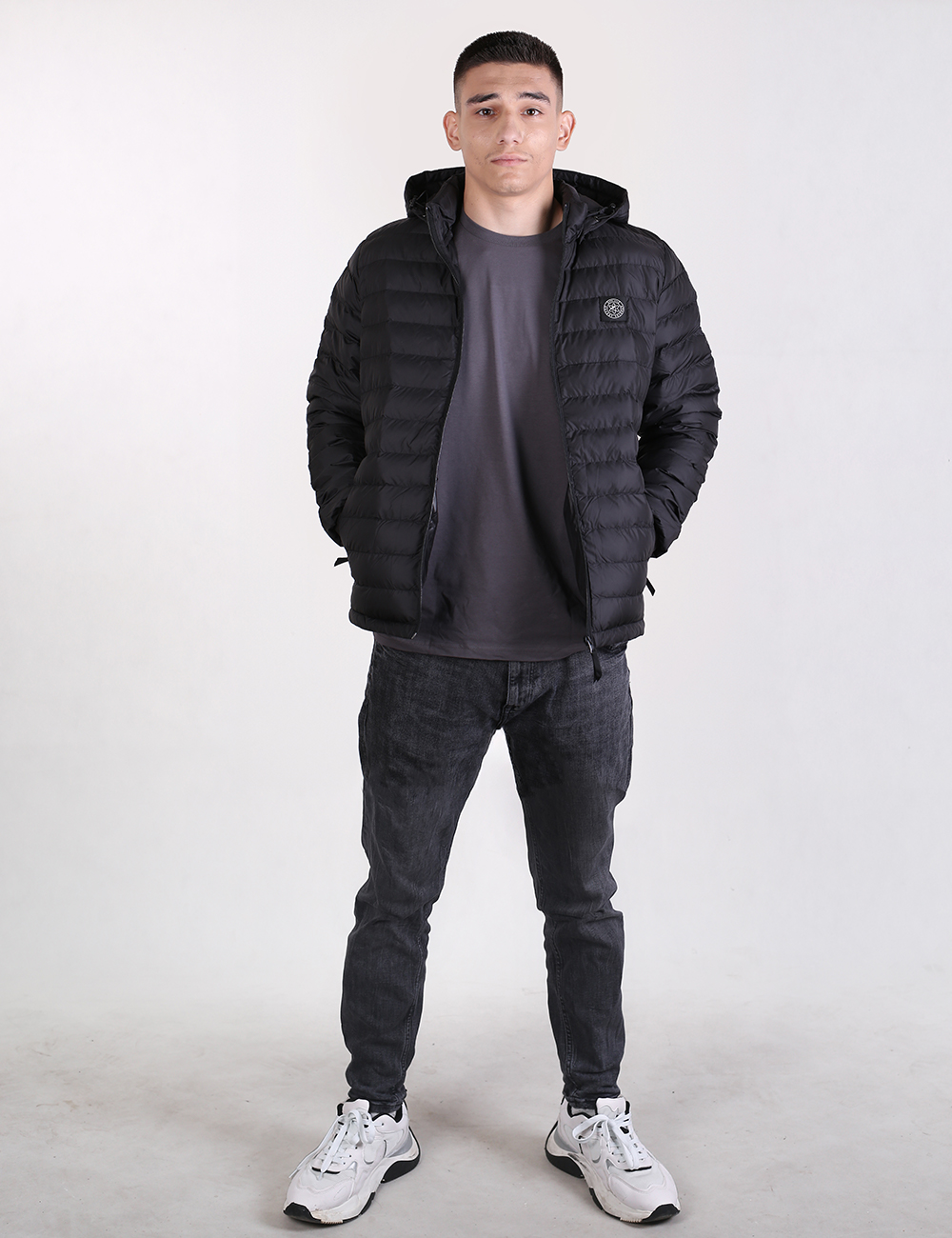 A young man stands facing forward against a plain light gray background. He is wearing a black hooded puffer jacket, which is left unzipped over a dark gray t-shirt. He is also wearing charcoal gray slim-fit jeans and chunky white sneakers with dark accents. His hands are tucked into his jacket pockets, and he has a neutral expression.
