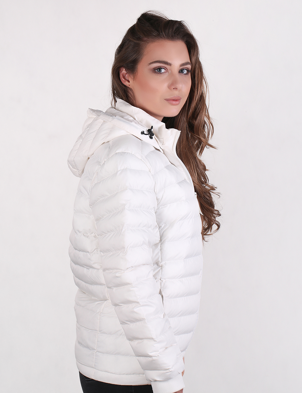 A woman wearing a white puffer jacket with a hood, shown in profile against a plain light gray studio background.