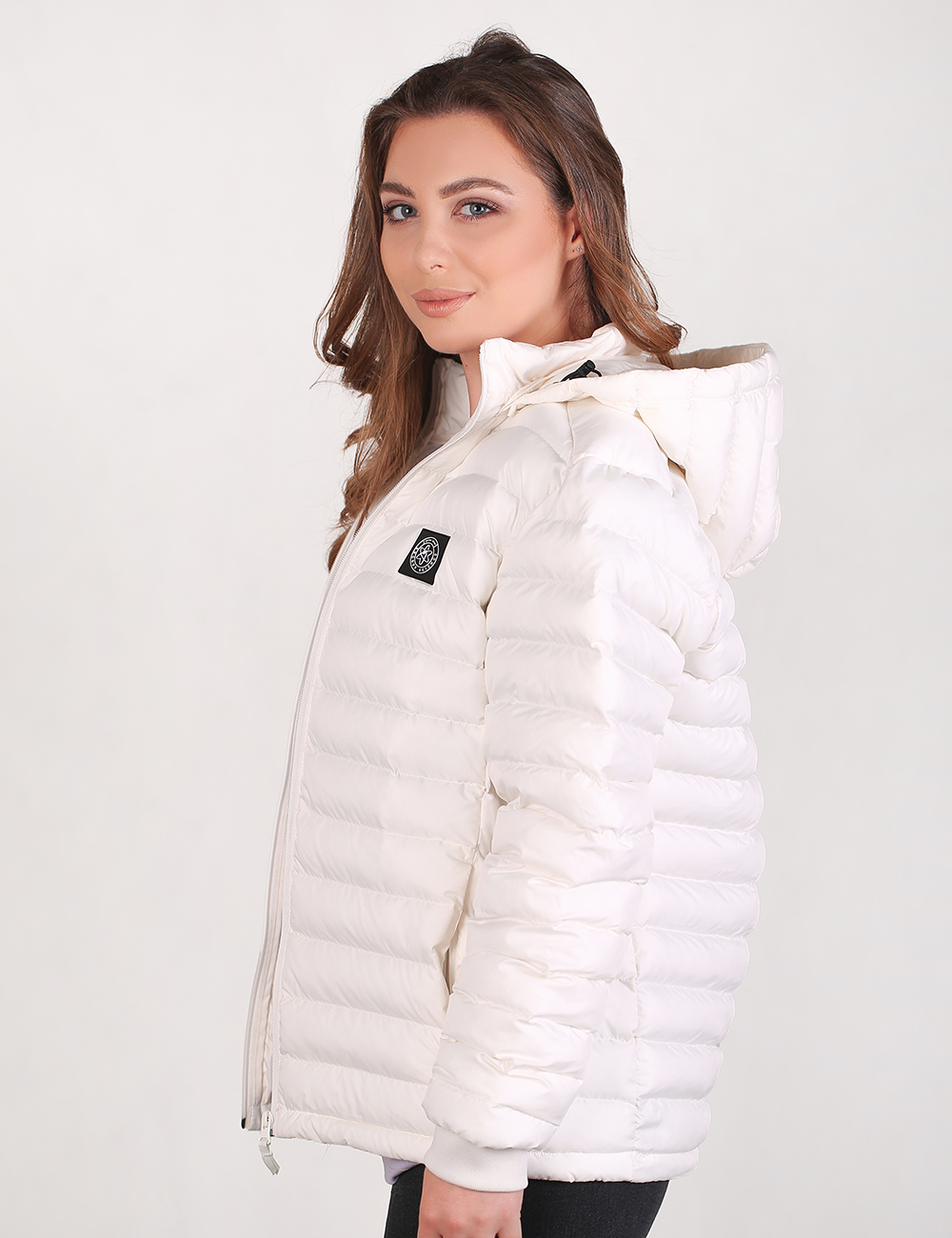 A woman wearing a white puffer jacket with horizontal quilting and a hood, shown from a side profile against a plain light gray studio background.