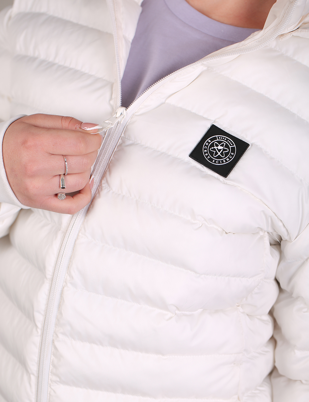 Close-up of a white puffer jacket with horizontal quilting, featuring a black square chest patch with a white circular logo. A hand with rings is visible near the zipper, worn over a light purple shirt.