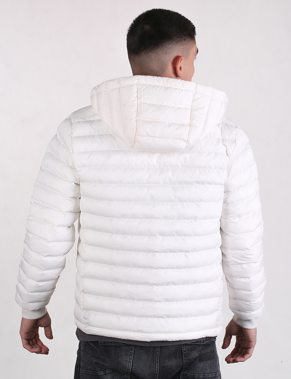 A man viewed from behind wearing a white hooded puffer jacket with horizontal quilting, paired with dark gray jeans, against a plain light gray studio background.