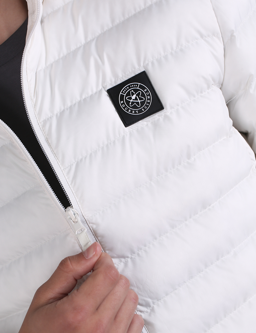 Close-up of a white puffer jacket with a black square chest patch featuring a circular logo, with a hand pulling the front zipper.