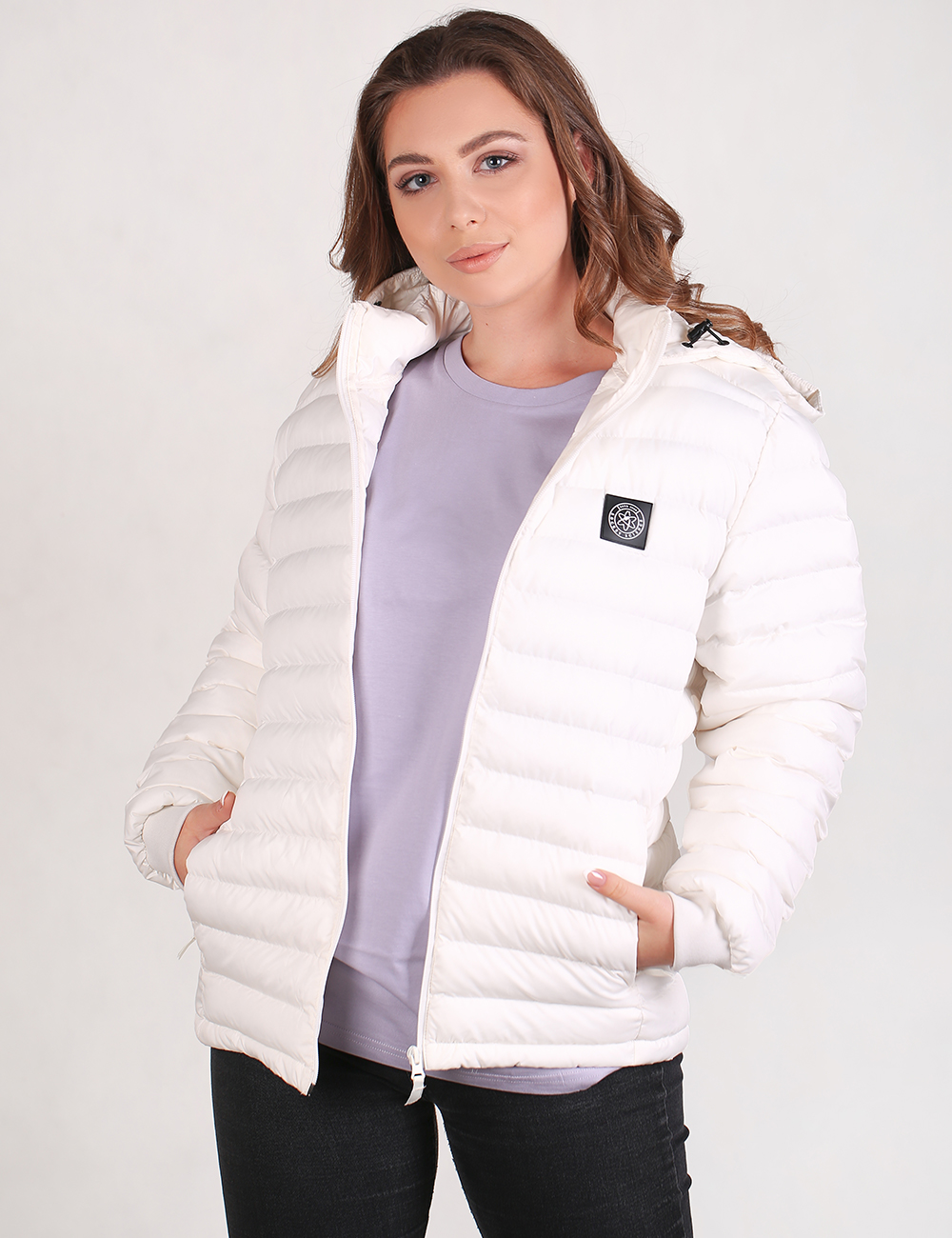 A woman wearing a white puffer jacket over a light purple sweatshirt and black pants, standing against a plain light gray studio background.