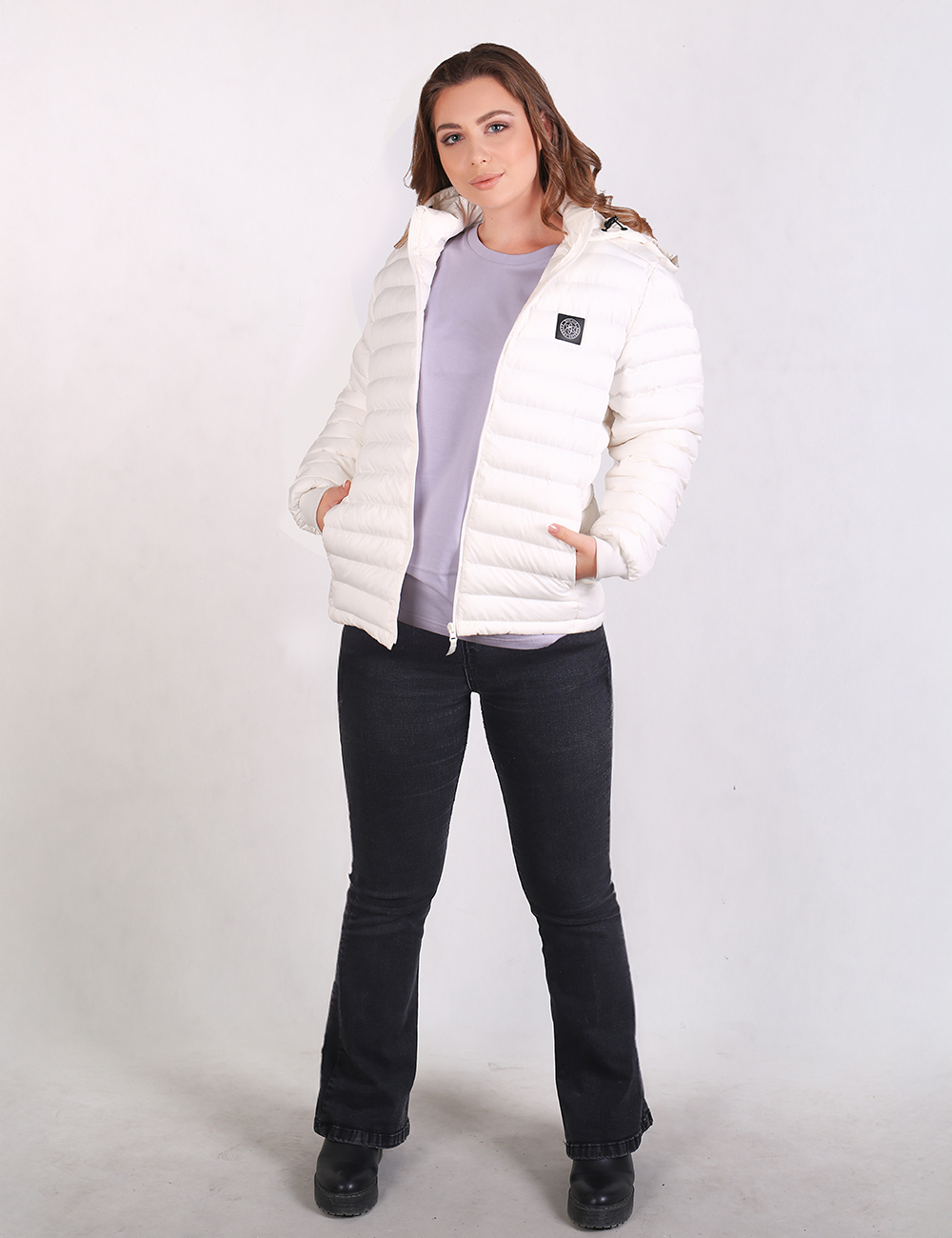 A woman modeling a white puffer jacket over a light purple top and black jeans, standing against a plain light gray studio background.