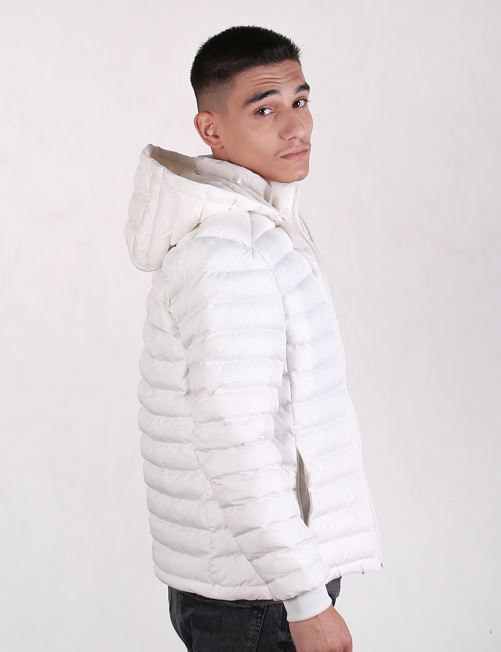 A young man wearing a white puffer jacket with a high padded collar and hood, shown in a side profile against a plain light gray studio background.