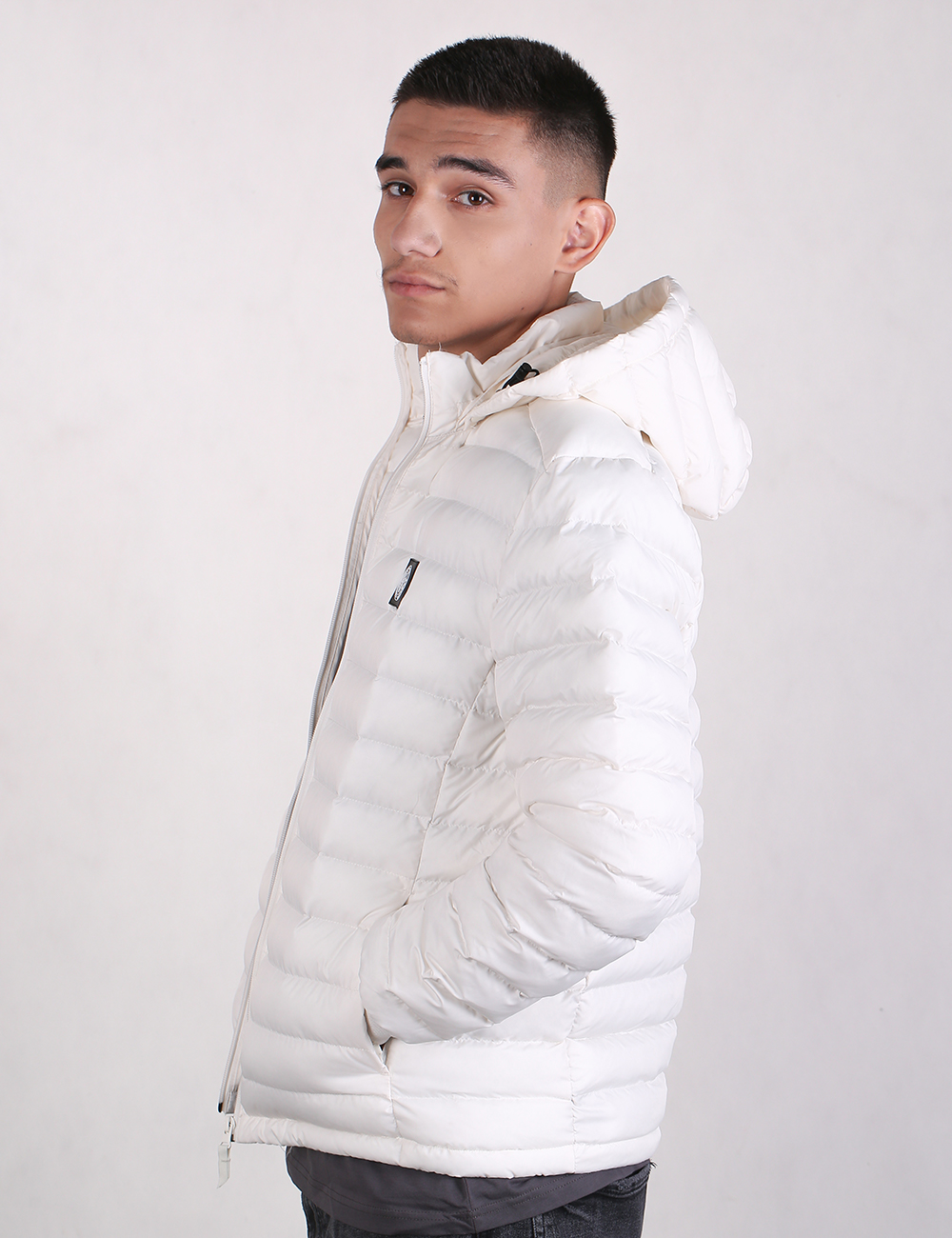A young man wearing a white hooded puffer jacket, shown in profile against a plain light gray studio background.