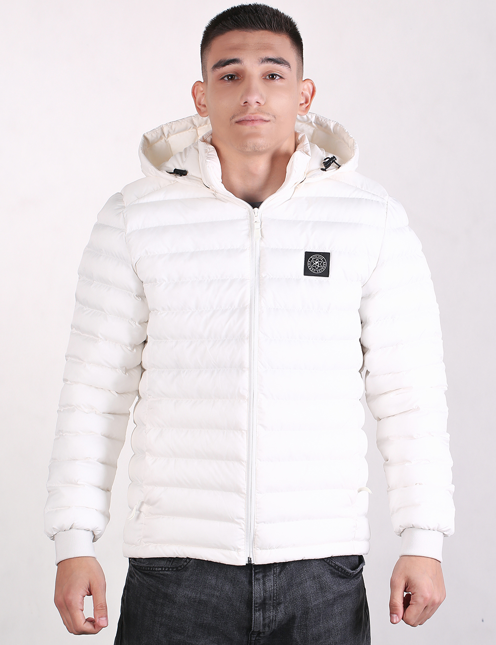 A man wearing a white puffer jacket with a zip-up front, hood, and a small black logo patch on the left chest, paired with dark gray jeans, against a plain white background.