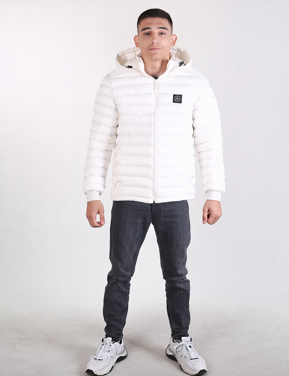 A man wearing a white puffer jacket with a faux-fur trimmed hood, dark gray jeans, and white sneakers, standing against a light gray studio background.