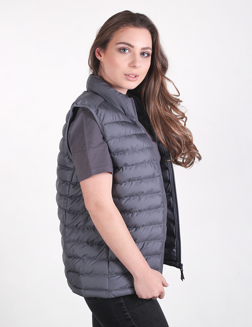 A woman with long, wavy brown hair posing from a side profile against a plain white background. She is wearing a dark grey, quilted puffer vest with a high collar over a dark grey short-sleeved t-shirt and black jeans.