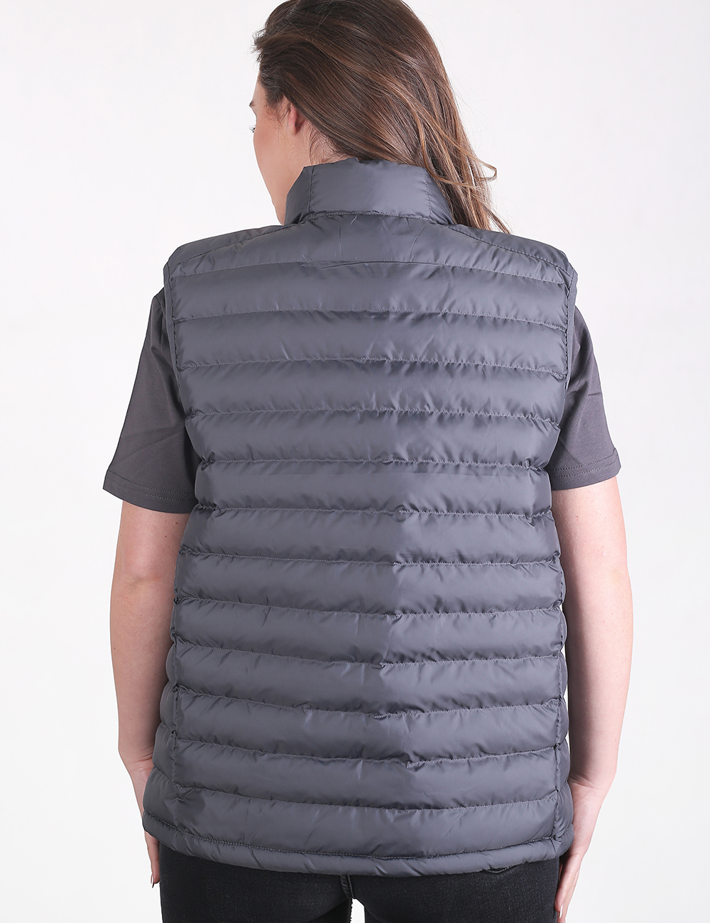 A rear view of a person with long brown hair wearing a dark charcoal grey puffer vest over a short-sleeved grey t-shirt and black pants. The vest features horizontal quilted stitching and a high, padded collar.