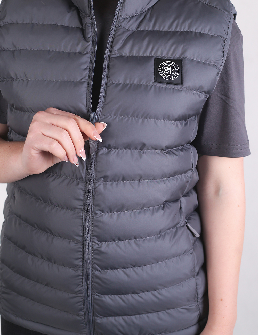 A close-up shot focuses on a person wearing a dark gray puffer vest over a matching gray short-sleeved t-shirt. The vest features horizontal quilted baffles and a front zipper, which the person is gripping with their left hand as if adjusting it. On the left chest area, there is a small, rectangular black patch containing a circular graphic logo.