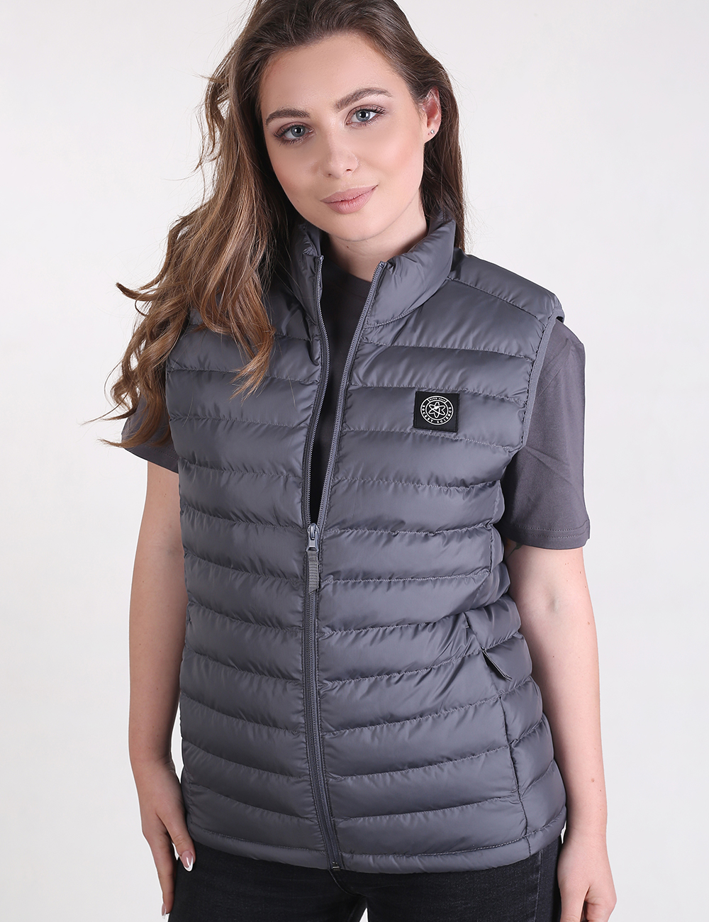 A young woman with long, wavy brown hair is posing while wearing a dark grey, quilted puffer vest. The vest is zipped up to her neck and features a small, round patch on the left side of the chest. Underneath, she wears a matching grey short-sleeved t-shirt and dark pants. The background is a solid, light grey.