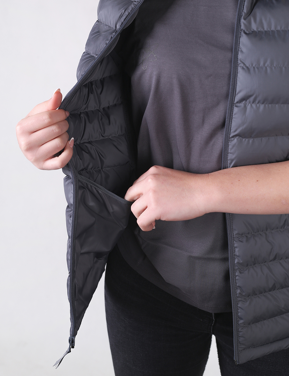 A close-up shot of a person wearing a dark grey quilted puffer vest over a matching grey t-shirt and black pants. The person's right hand is holding the open front flap of the vest to reveal a deep, interior drop-in pocket, while their left hand is partially tucked inside it.