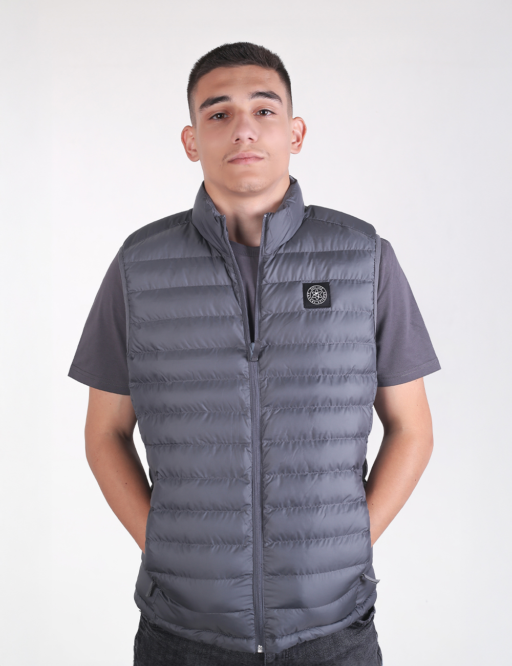 A young man looking directly at the camera while wearing a deep metal grey padded vest over a matching grey short-sleeve t-shirt.