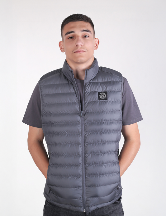 A young man looking directly at the camera while wearing a deep metal grey padded vest over a matching grey short-sleeve t-shirt.
