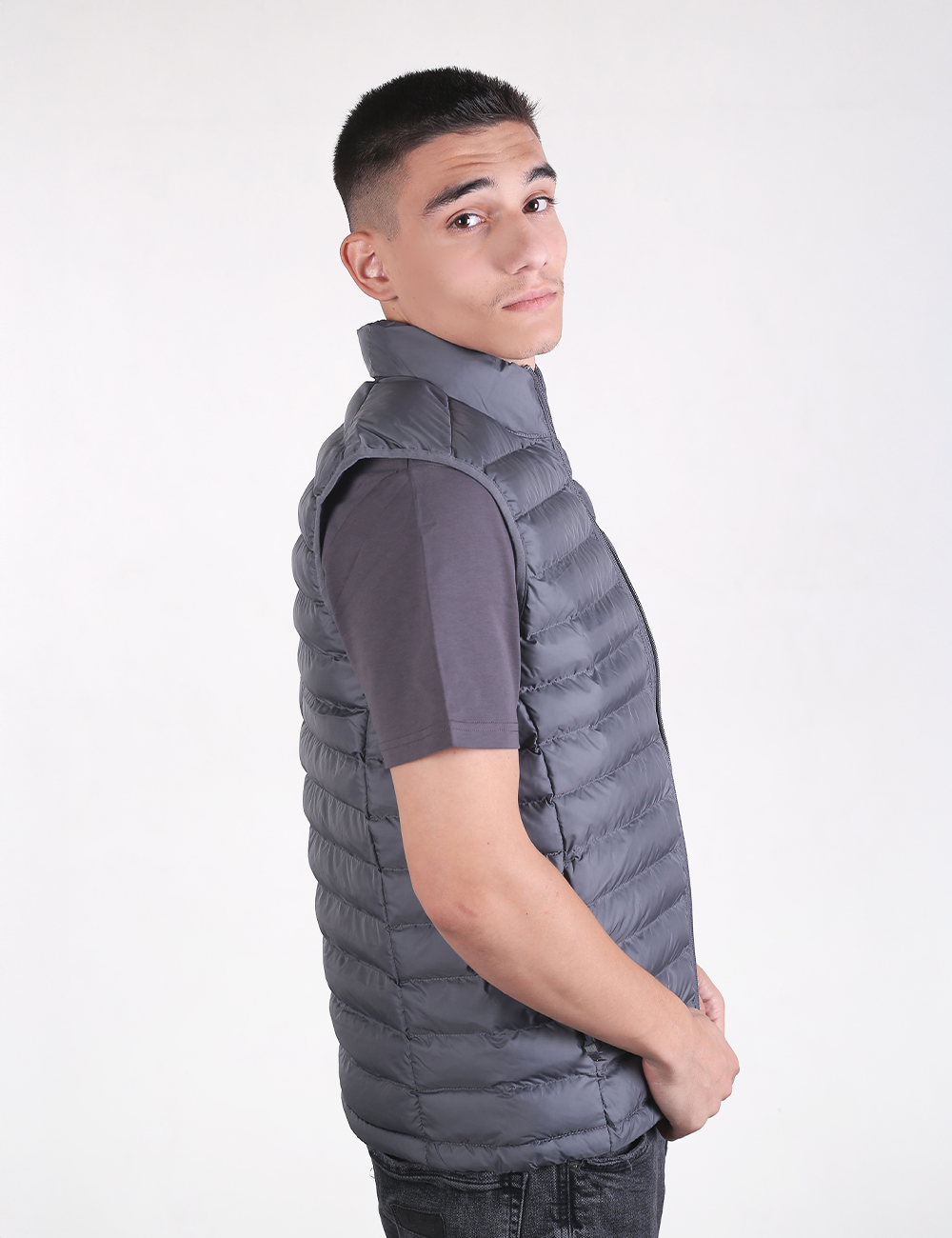 A young man with short dark hair looking over his right shoulder. He is wearing a dark grey, horizontally quilted puffer vest over a plain, dark grey short-sleeved t-shirt. The background is a solid, light off-white color.