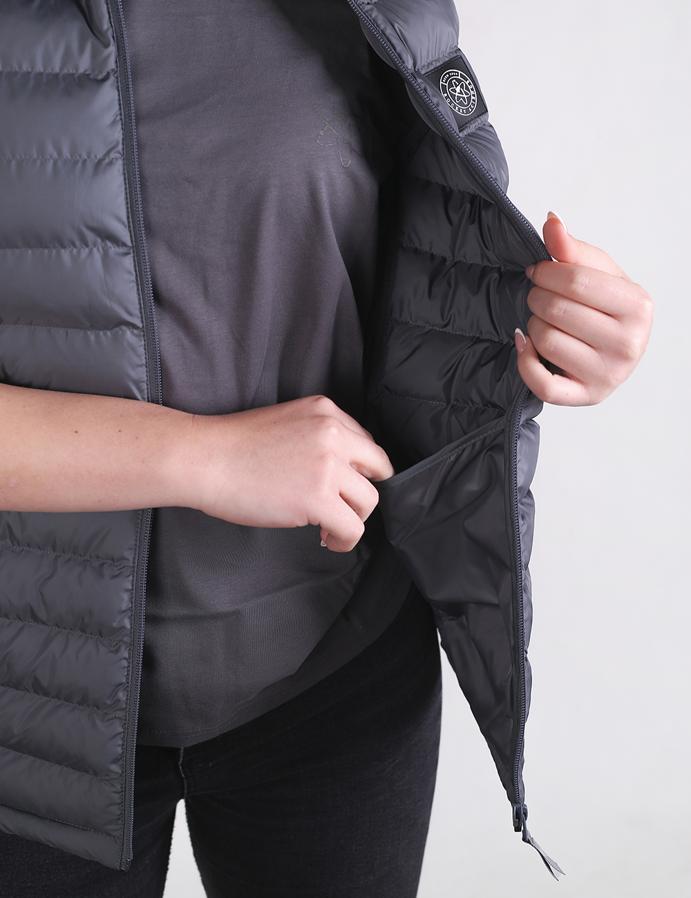 A close-up shot focuses on a person wearing a dark gray puffer vest over a matching gray short-sleeved t-shirt. The vest features horizontal quilted baffles and a front zipper, which the person is gripping with their left hand as if adjusting it. On the left chest area, there is a small, rectangular black patch containing a circular graphic logo.