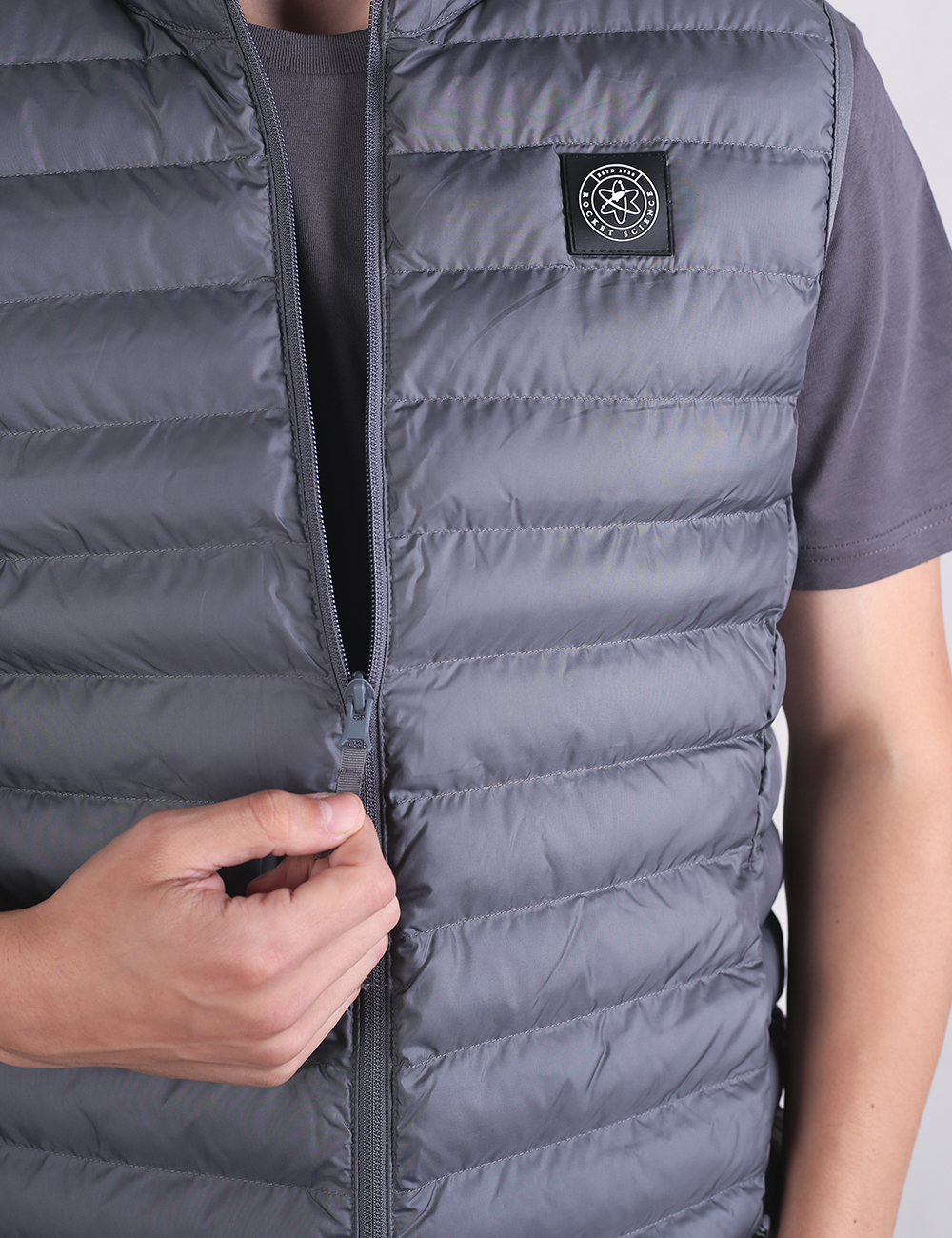 A person wearing a sleeveless, grey padded vest over a darker grey t-shirt is shown from the chest down. The person's left arm is relaxed at their side, while their right hand is adjusting the zipper of the vest near the center. The vest features thin, horizontal quilted panels and a small, circular black patch logo on the left chest.
