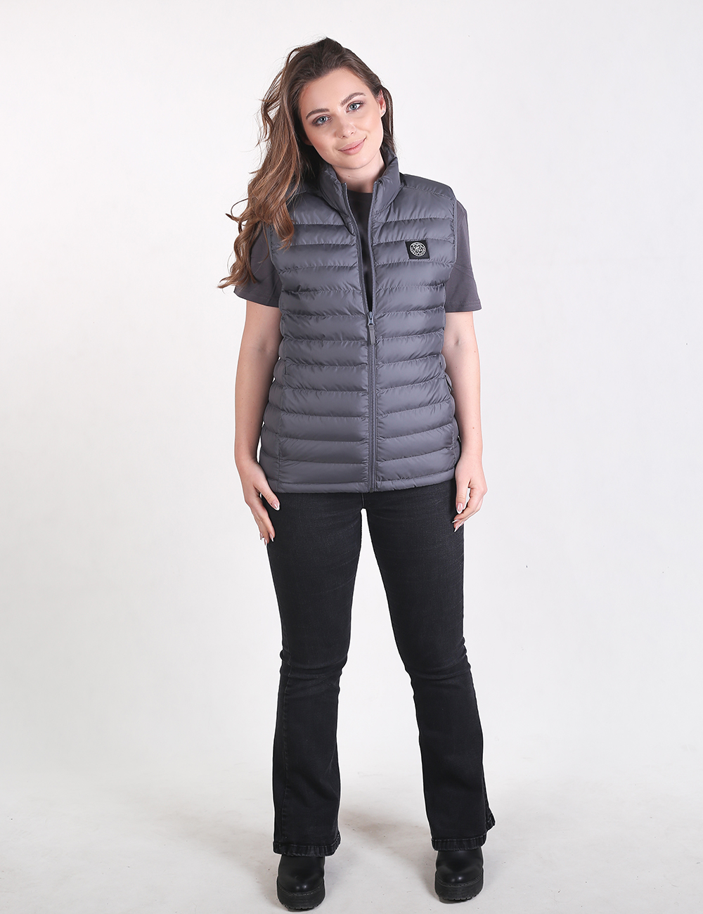 A woman with long brown hair poses against a white background, wearing a dark grey quilted puffer vest, a grey short-sleeved t-shirt, black flare jeans, and black shoes.