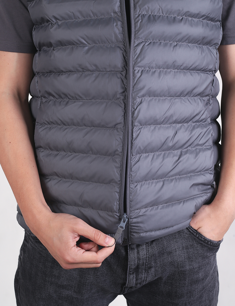 A person wearing a sleeveless, grey padded vest over a darker grey t-shirt is shown from the chest down. The person's left arm is relaxed at their side, while their right hand is adjusting the zipper of the vest near the center. The vest features thin, horizontal quilted panels and a small, circular black patch logo on the left chest.