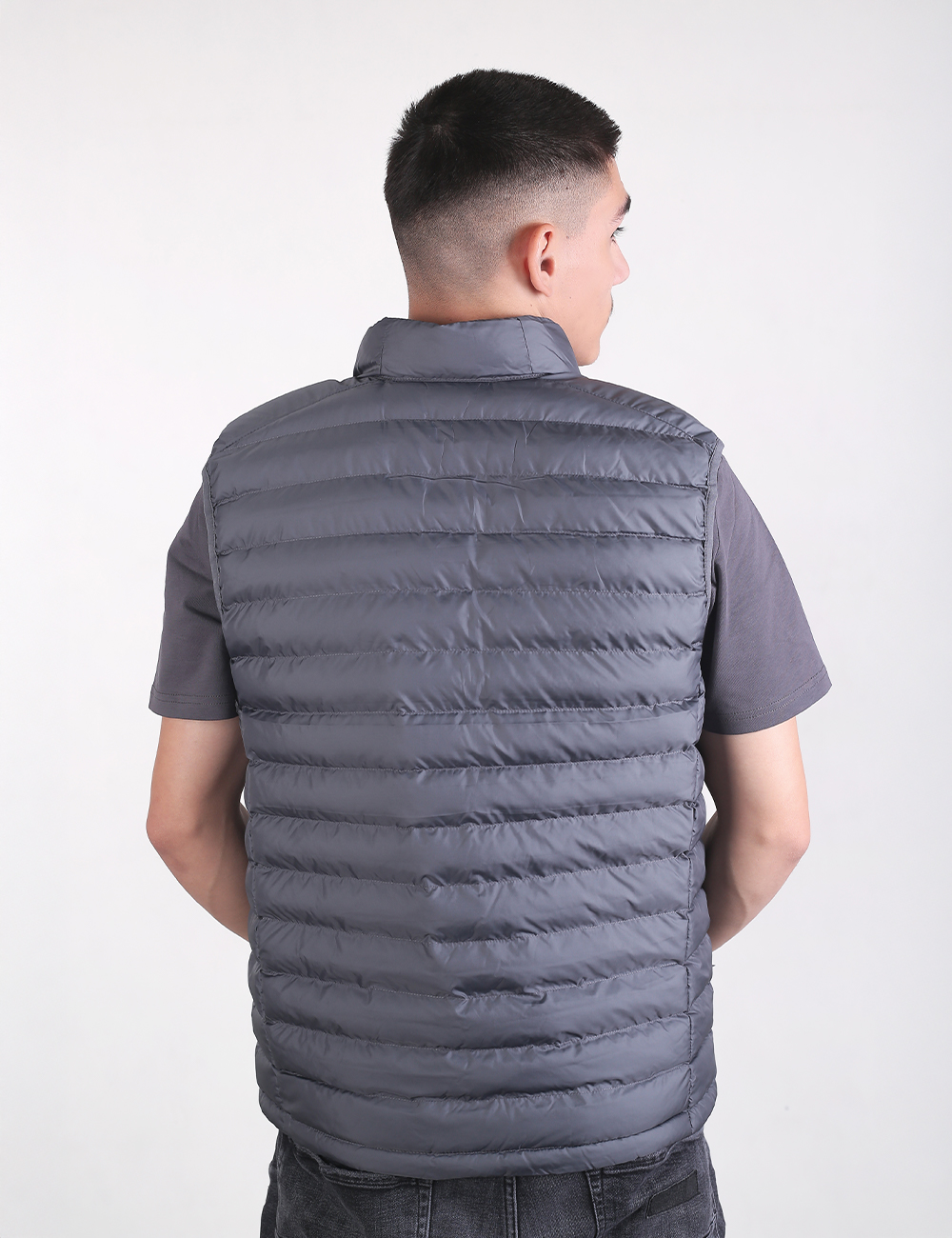A man viewed from behind from the waist up, wearing a dark grey, horizontal-quilted puffer vest over a matching short-sleeved grey t-shirt. He has a short fade haircut and is looking slightly over his right shoulder against a plain, light grey background.