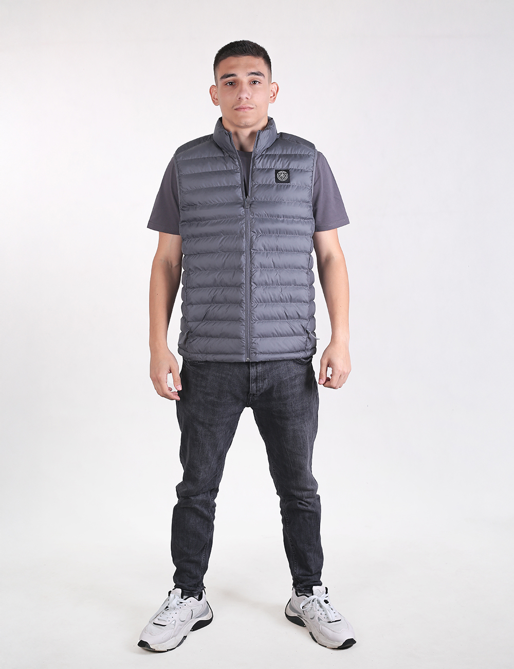 A man standing against a white background, wearing a grey padded puffer vest with a circular logo on the left chest, a grey short-sleeve t-shirt underneath, dark grey or black slim-fit jeans, and white sneakers with black accents.