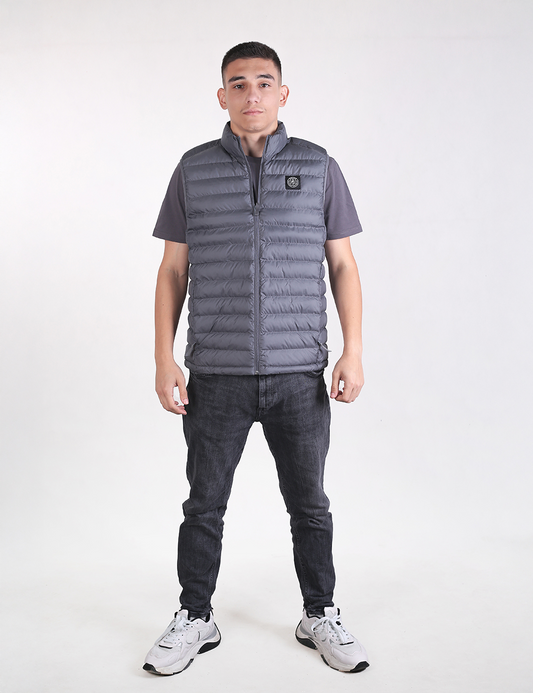 A man standing against a white background, wearing a grey padded puffer vest with a circular logo on the left chest, a grey short-sleeve t-shirt underneath, dark grey or black slim-fit jeans, and white sneakers with black accents.
