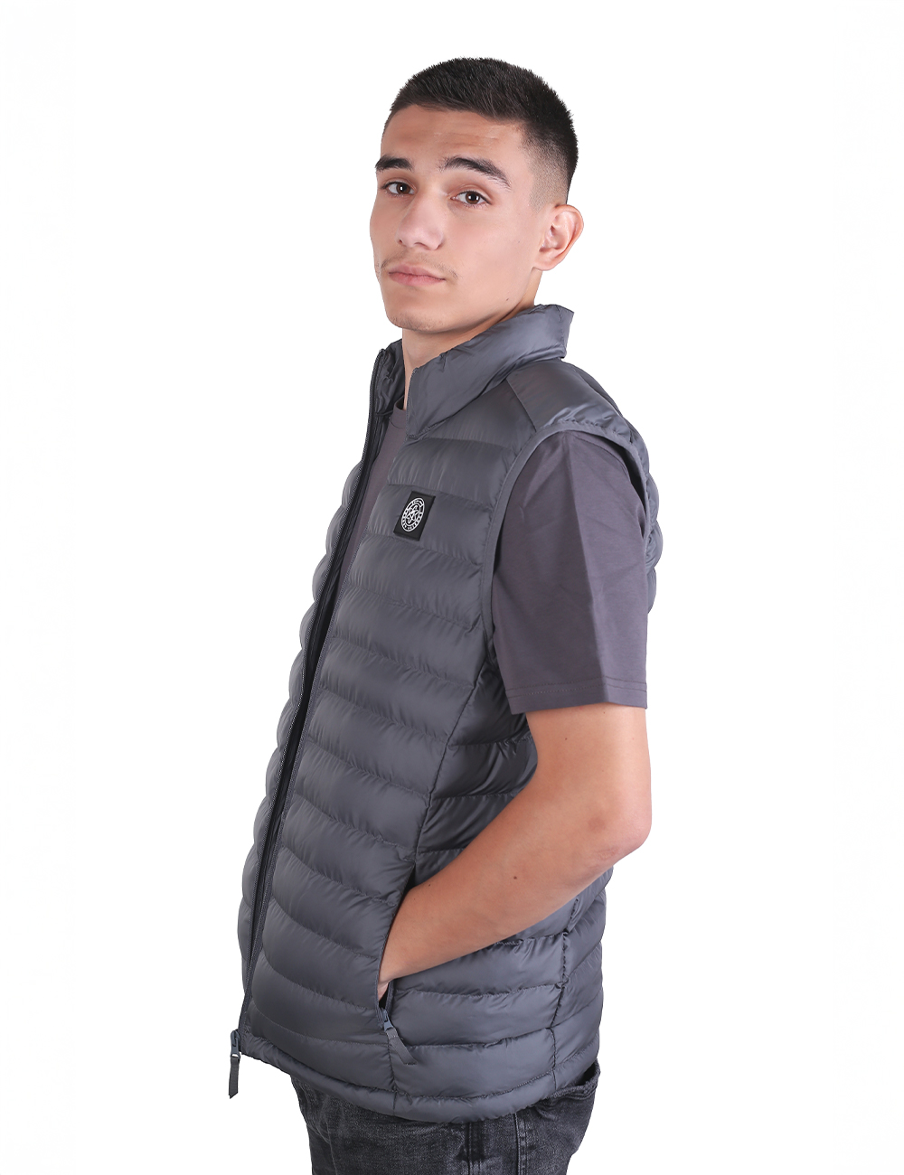 A young man with short dark hair, standing in profile while looking toward the camera against a plain white background. He is wearing a dark grey, sleeveless padded puffer gilet vest over a matching grey short-sleeved t-shirt. His left hand is tucked into the side pocket of the vest, and he wears dark jeans.