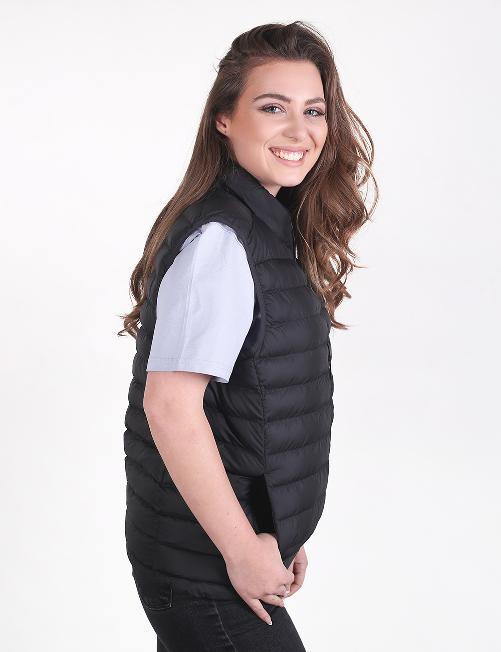 A woman with long brown hair stands sideways, smiling warmly while wearing a sleeveless black quilted puffer vest over a simple white t-shirt and dark pants.
